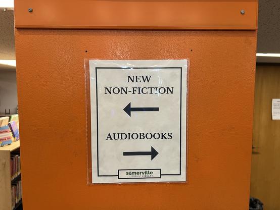 A sign hung on the end of a bookcase in the Somerville Public Library. To the left is new non-fiction, to the right is audiobooks.