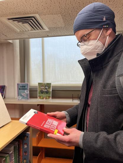 I’m standing in the library while wearing an N95 mask, a blue winter cap, and a long black coat. I’m looking down at my hands while holding a copy of my latest book, YOU DESERVE A TECH UNION, which my local library apparently just procured.