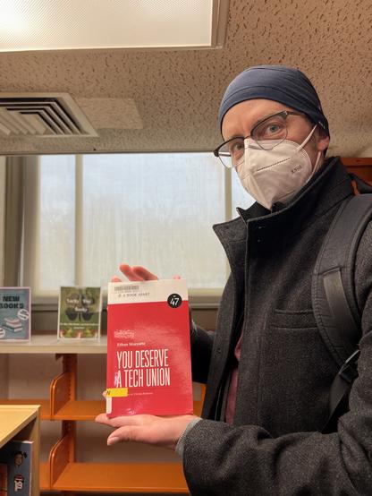 I’m standing in the library while wearing a KN95 mask, a blue winter cap, and a long black coat. I’m looking at the camera while I hold up a copy of my latest book, YOU DESERVE A TECH UNION, which my local library apparently just procured.