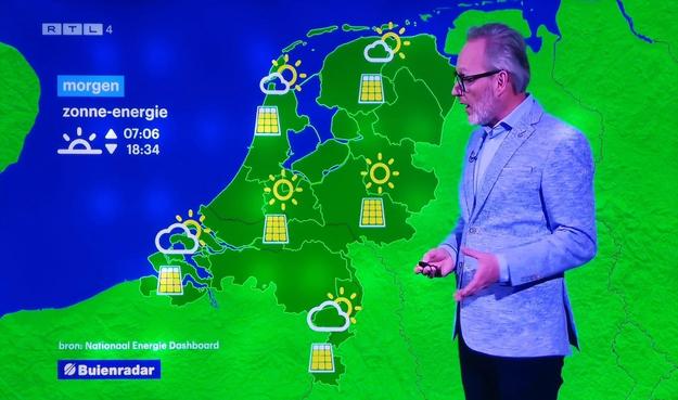 Television weather forecast showing a map with solar energy symbols indicating sunny conditions, with a weather presenter standing beside it.