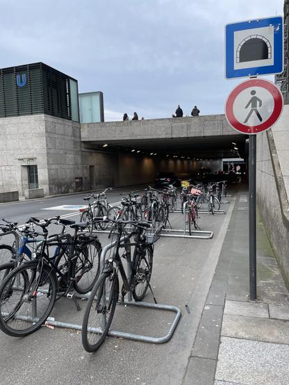 Fahrradständer vor einem Tunnel, rechts ein Schild dem gemäß der Durchgang für Fußgänger verboten ist