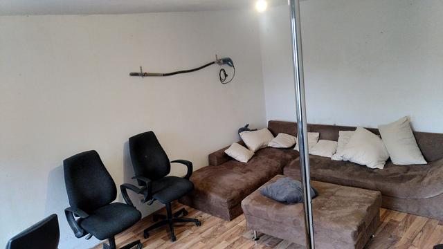 Ein kleiner renovierungsbedürftiger Raum mit einem großen braunen Eck-Sofa mit beigen Kissen. Links daneben stehen schwarze Bürostühle. Im Vordergrund steht eine verchromte Poledance-Stange. An der Wand hängt ein 2 Meter langer flexibler Schlauch auf 2 Haken. Das Eine Ende ist mit einem Metallzylinder abgeschlossen und das andere Ende ist an eine Maachine montiert, die wie eine Bohrmaschine aussieht (nicht im Bild erkennbar: auf der Maschine steht "Vibrator 1300W")