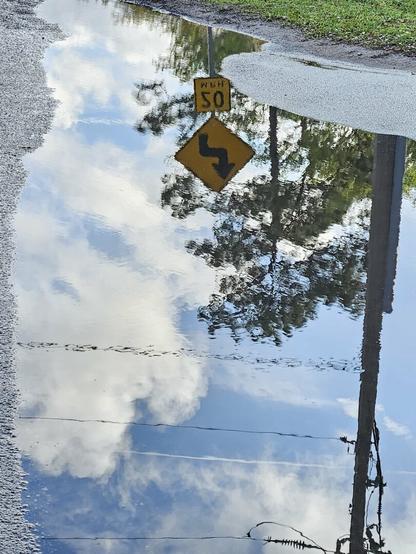 Large rain puddle on side of a roadway, with nearby traffic signage and power lines reflecting inverse images with surrounding sky and cloud formations.