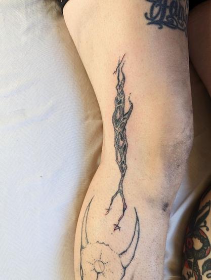 This image is of the right leg of a person with somewhat fair skin. Two other tattoos are somewhat visible in the frame, but the focus is of a stylized segment of a cholla (a kind of a cactus) skeleton, which looks a bit like a branch fragment with a pattern of holes throughout. The tattooer has extended the branch in a stylized way, trailing the terminal ends of it into simple sigils comprised of intersecting lines and dots. The tattoo has been hand poked, so the shading is dotwork.
