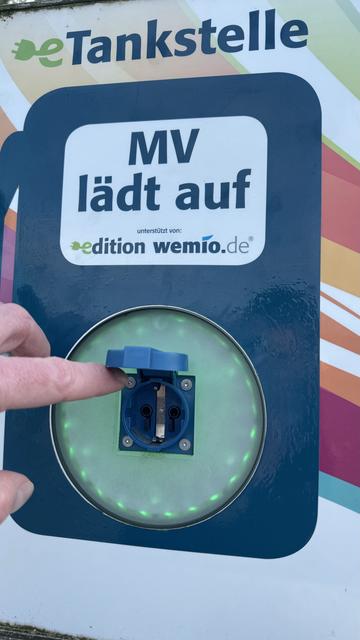 Der Finger einer Person zeigt auf eine Schuko-Steckdose mit der Aufschrift "eTankstelle MV lädt auf", umgeben von einem leuchtenden grünen Licht.