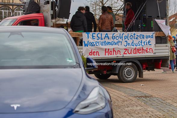 Ein Auto von Tesla steht auf einem Parkplatz, dahinter ein Lautsprecherwagen mit einem Banner: "Tesla, Golfplätzen und wasserintensiver Industrie den Hahn zudrehen".