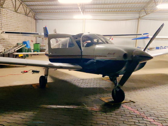 A blue-and-white Piper Archer III with open door sitting at night in a hangar lit by fluorescent tubes. A small ground-power unit with cable is lying on the ground beside it. Parts of other light aircraft, ladders, chairs and other things typical for a hangar can be seen in the background.