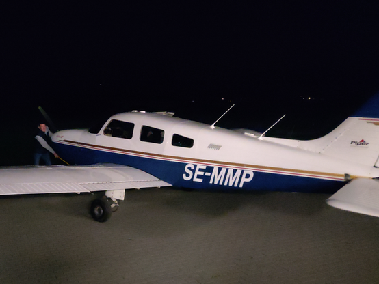 A blue-and-white Piper Archer III on the ground at night, the background is almost completely black, diffuse white light is illuminating the plane and one person is pulling it by the nose gear with a towbar.
