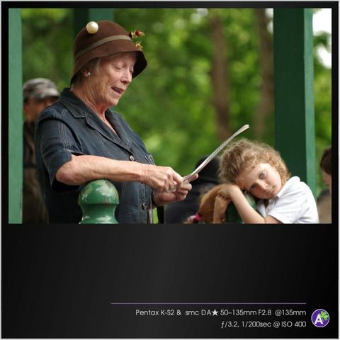 a color outdoor photograph. an older woman stands on a porch or in a gazebo. she is reading from a laminated page. her face appears animated and energetic. several people listen in the background. one small girl in the background looks very bored and restless. she climbs on the fence railing and looks away. there are no animals or vehicles in this picture.