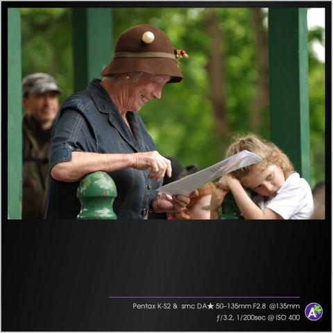 a color outdoor photograph. an older woman stands on a porch or in a gazebo. she is reading from a laminated page. her face appears animated and energetic. several people listen in the background. two young children, a boy and a girl, look very bored. the boy climbs a pole while the girl hides her head in her arms. there are no animals or vehicles in this picture.