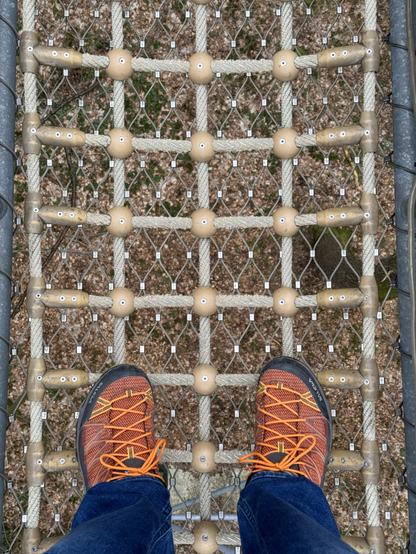 Das Bild ist eine Draufsicht auf eine Person, die auf einer Seilbrücke steht und orangefarbene Schnürschuhe und Jeans trägt.