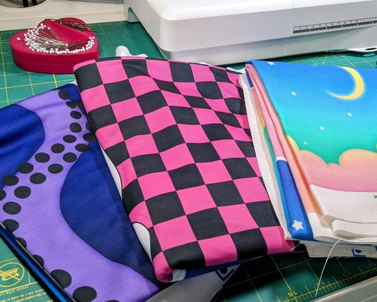 three folded yards of fabric, featuring a purple tentacle design, a pink and black checkerboard print, and a pastel moonscape design