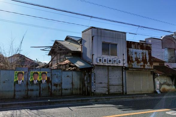 From right to left: a number of small square looking two-storey buildings side by side. They used to be business but all the shutters are closed. Then there is a fence and behind it a glimpse of a building that used to be there. Some debris still sticks to the shared wall with the neighbor. On the fence somebody pasted the candidate poster for the local election. The sun is coming in hard from the side. Nobody there. All very gloomy.