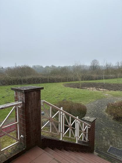 Ein Blick von einem Balkon auf einen Garten und ein Feld mit Bäumen in der Ferne an einem bewölkten Tag.