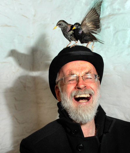 Terry Pratchett laughing heartily while two starlings sit atop his hat