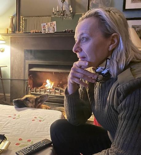 An older white woman with Predator blonde hair and a dubious but warm military-style khaki jumper sits near a table which itself is near a lit fireplace. Indoors in a cosy room, a German Shepherd rests her muzzle on the table top, herself nearest the fireplace. Both are looking out of the frame, the glow from the TV on the woman's face.