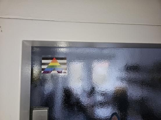 Der obere linke Teil einer gläsernen Bürotür ist zu sehen. Darauf klebt die Straight Ally Fahne: ein Regenbogen-A auf schwarz-weißen Streifen.
