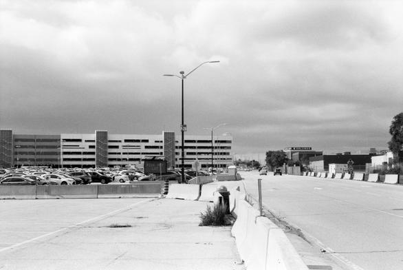 A three lane roadway passes an expansive surface parking lot on its way to a larger multilevel parking lot.