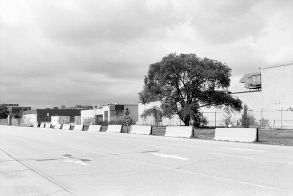 Three lanes pass concrete bollards and low industrial buildings.
