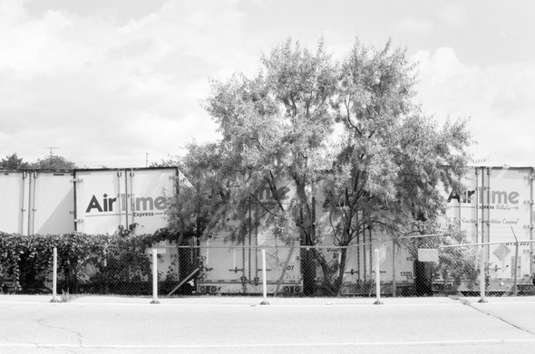 A veritable wall of tractor-trailers backed against a fence with one tree wedged in somehow. The trailers are labelled “AirTime Express”.