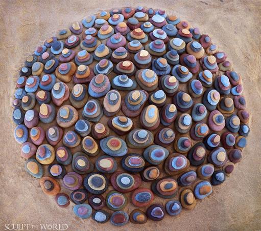 A circle of rocks from the seashore. The larger rocks are at the bottom. Carefully stacked on top are a series of smaller stones. Each stone is a distinct colour from the others in its stack.