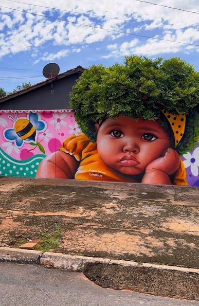 Streetartwall. The mural of a little girl whose hair consists of a green tree was sprayed/painted on a long street wall. The head of a little black girl in a yellow dress has been sprayed on a wall with a large conifer or shrub behind it. The bush grows over the wall and forms the child's wild mop of hair. The girl rests her head on one arm and looks sullenly in the direction of the viewer. Next to the child, a fat bee with blue wings flies over a turquoise-pink wall of flowers.