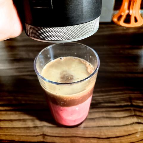 Espresso being poured into a glass of pink blood orange juice on a wooden surface.