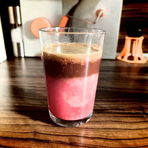 Layered beverage with pink blood orange juice and brown espresso layers in a glass on a wooden surface, with kitchen utensils in the background.