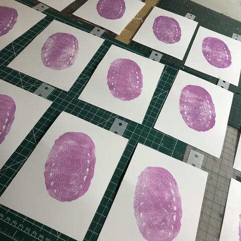 A grid of drying linocut prints on a tabletop with cutting mats. There are at least 13 prints in the photo and they are on white printmaking paper 4” x 6”. They each have one registration tab attached and show the first stage of my chiton print with the cool pale pink ovoid shape with some white triangles and fine striped lines on the plates.