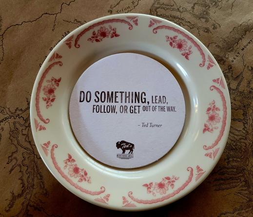 Color Photo
An old brown map is the base of the photo. The map appears to be of the Yellowstone area in the United States. 

A white round saucer with pinkish red decorative flourish around the edge called transferware. 

A coaster sits inside the saucer with this text:

Do something, lead, follow, or get out of the way. -Ted Turner