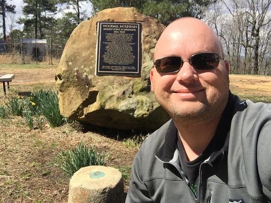 Man taking a selfie with a commemorative plaque on a large rock, detailing Woodall Mountain as the highest point in Mississippi.