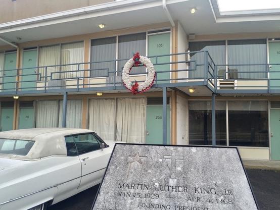 A historical site with a memorial plaque for Martin Luther King Jr. in the foreground, a vintage white car, and a motel balcony with a wreath marking a significant location.