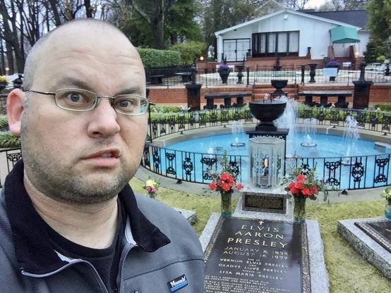 A person taking a selfie in front of Elvis Presley's grave at Graceland, with a fountain in the background.