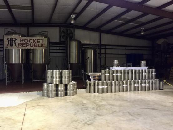 Brewery interior with large fermentation tanks and stacked metal beer kegs.