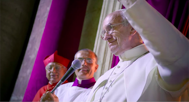 An elderly man in a white papal shirt and jacket waving while standing next to a microphone.