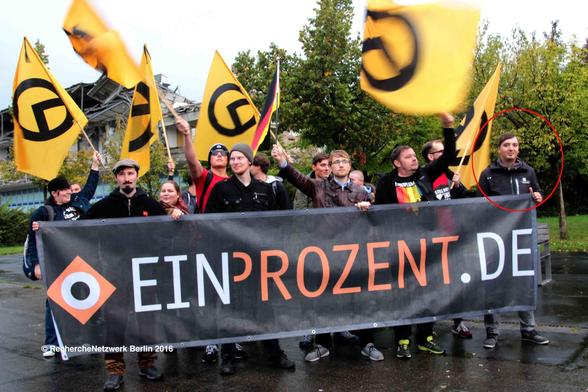 Eine Gruppe von ca. 10 Personen steht im Regen hinter einem Banner von "EinProzent". Es sind sechs gelb-schwarze Flaggen der Identitären und eine BRD-Flagge zu sehen. Links am Banner steht Konrad Kohlhas aus Eisenach, mittig steht Marcel Waschek aus Jena. Beide sind Mitgründer der Thüringer Identitären. Rechts am Banner steht rot eingekreist Christoph Walther.