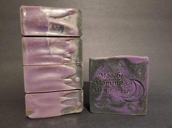 On the Left are found black and purple soaps stacked on their side to show the top detail. On the Right is a single bar of black and purple swirled soap with "Moody Momma's Menagerie" and a crescent moon stamped