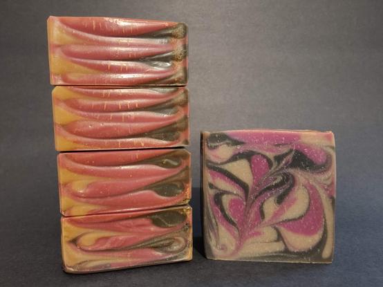 On the Left four soaps are stacked on their side to show the top detail. On the Right is a single bar of black, raspberry and cream soap showing the side with "Moody Momma's Menagerie" and a crescent moon stamped on the bar