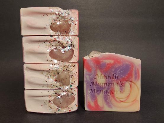 On the Left are four bars or soap stacked on their side to show the top detail with biodegradable glitter and a Rose Quartz tumble on each bar. On the Right is a single bar of the pink, purple and cream colored soap with "Moody Momma's Menagerie" and crescent moon stamped
