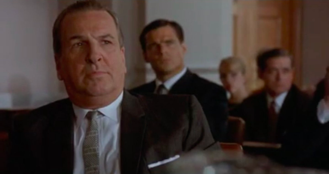 A courtroom scene showing an older man in a suit sitting at a defendant's table, looking up at the judge and jury.
