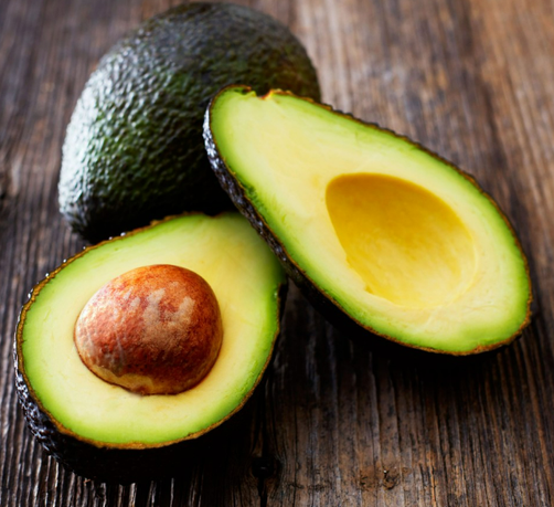 Avocados, whole and split. The center pit is still in place in one half of one. They sit on a wooden table.