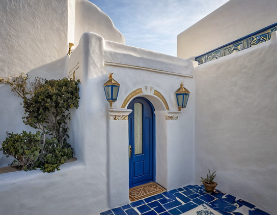 White Mediterranean villa detail with a contrasting vivid blue door, golden lanterns, and geometric tiles in sunlight.