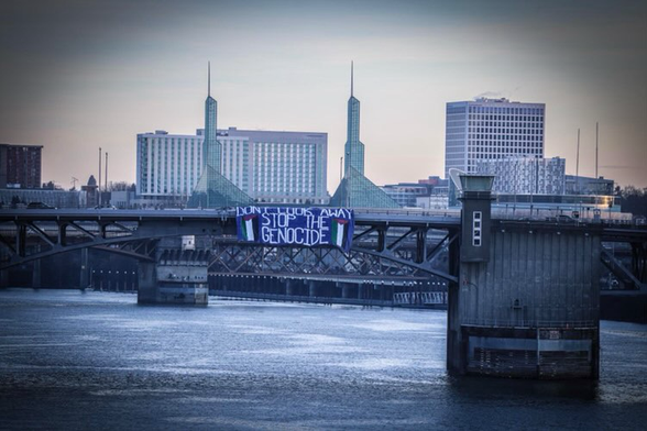 A massive banner hanging on the Morrison bridge with a video of Portland and the river. The banner is blue and the text says “don’t look away” “stop the genocide” on all capital white letters with two Palestinian flags on both bottom corners