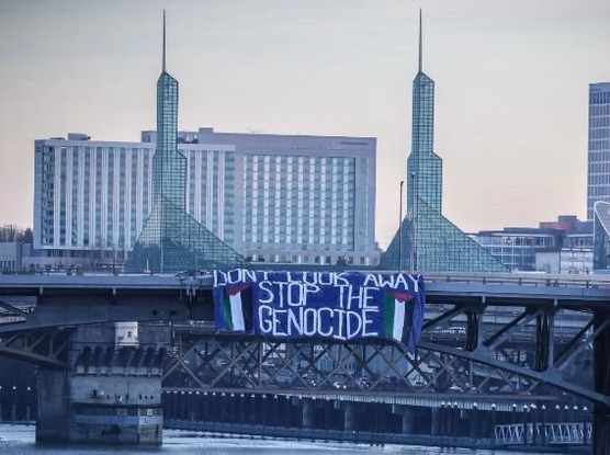A massive banner hanging on the Morrison bridge with a video of Portland and the river. The banner is blue and the text says “don’t look away” “stop the genocide” on all capital white letters with two Palestinian flags on both bottom corners