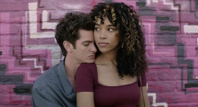 Andrew Garfield and Alexandra Shipp share an embrace. Behind them is a colorful painted brick wall with geometric designs.
