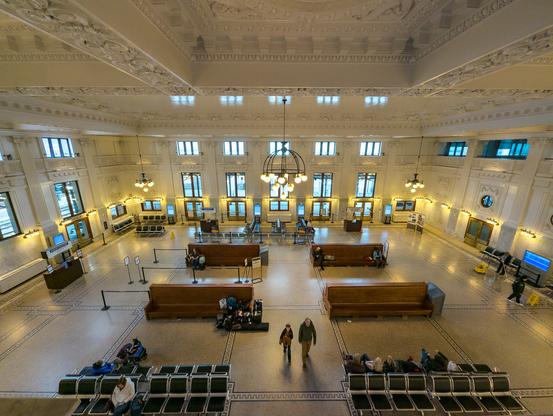 Interior view of the waiting room at King Street Station. Terrazzo flooring. Coffered ceiling with ornate design. Benches and passengers waiting with their luggage.