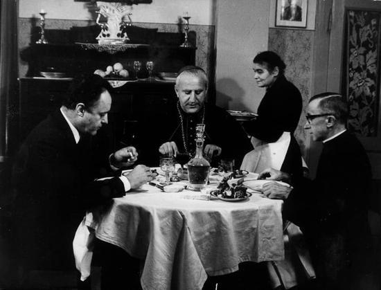 The image portrays a domestic scene with three individuals seated around a dining table. On the left, there is a person dressed in a black coat with a white shirt, wearing glasses and seemingly engaged in conversation or observing something on the table. In the center, another individual appears to be serving food from a bottle, contributing to the ambiance of a meal in progress. To the right, a third individual is depicted in mid-action, perhaps speaking or gesturing towards someone outside the frame.
The setting suggests an indoor domestic environment with a tablecloth and various dishes on the table. The attire of the individuals and the presence of items like cups and bottles indicate that this could be a formal dinner or gathering.
The image is black and white, which lends it a timeless quality. The style of clothing worn by the individuals suggests that the photograph might have been taken in the mid-20th century. There are no texts visible in the image to provide additional context or information about the event depicted or the identities of the individuals involved.