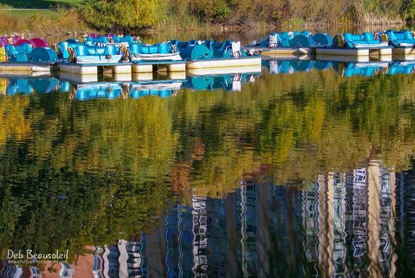 Turquoise and white paddle boats reflected on the water of a pond. Reflection of the trees and skyscrapers also visible.   Image at:  https://beautifulsunphotography.com/featured/paddle-boats-deb-beausoleil.html  See more art & blog at: https://beautifulsunphotography.com/ https://debbeausoleil.com