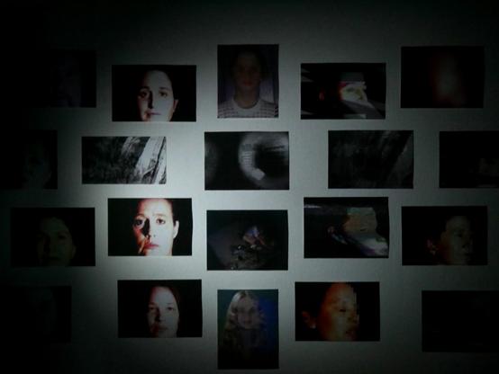 Farbfoto zeigt eine Wand in einem Ausstellungsraum, an der mehrere Fotos hängen, die meist Gesichter zeigen. Beleuchtet wird die Wand vom runden Lichtkegel einer Taschenlampe.