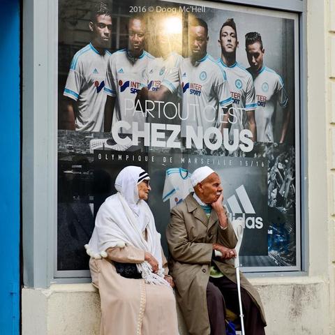 Un couple de vieux (peut-être né·es au sud de la Méditerranée) est assis adossé à une publicité de 2016 avec "Olympique de Marseille" juste au-dessus des deux têtes et pour slogan : "Partout c'est CHEZ NOUS".

(en)
An elderly couple (perhaps born south of the Mediterranean) sits against a 2016 advertisement with "Olympique de Marseille" just above their heads and the slogan: "Everywhere is HOME".